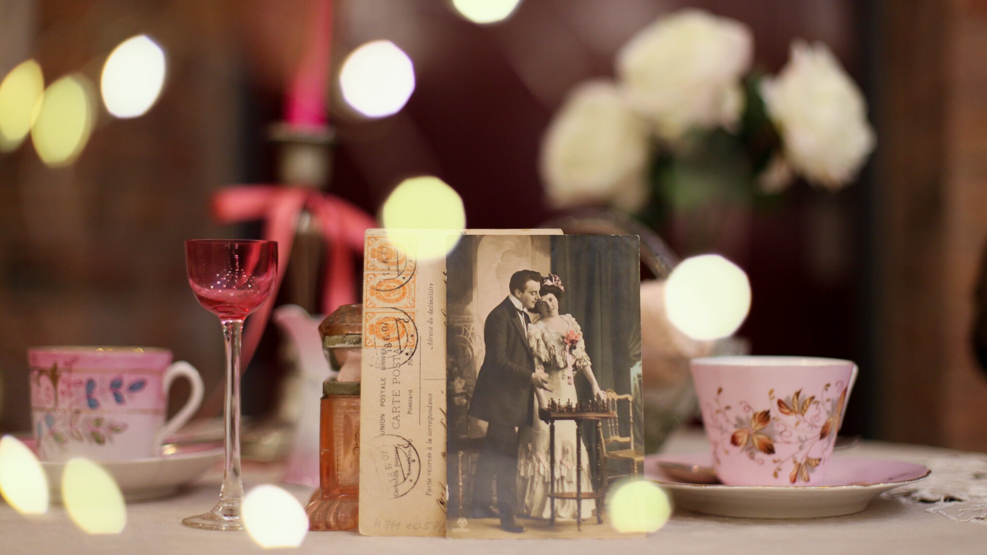 Romantisks kafijas galda servējums ar baltām rozēm, rozā kafijas krūzēm un liķiera glāzi, kā arī 20. gadsimta visu mīlētāju dienas atklātni centrā