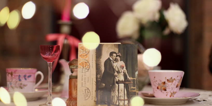 Romantisks kafijas galda servējums ar baltām rozēm, rozā kafijas krūzēm un liķiera glāzi, kā arī 20. gadsimta visu mīlētāju dienas atklātni centrā